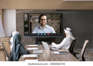 Videoconference talk between multicultural business teammates in conference room. Arabian businesspeople, entrepreneurs dressed in traditional Muslim wear having online meeting on tv monitor screen