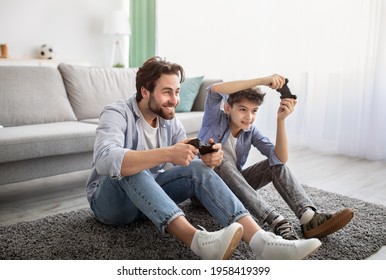 Videogames concept. Joyful father and son competing in online games, holding joysticks while sitting on carpet in living room and spending time together on weekend, copy space