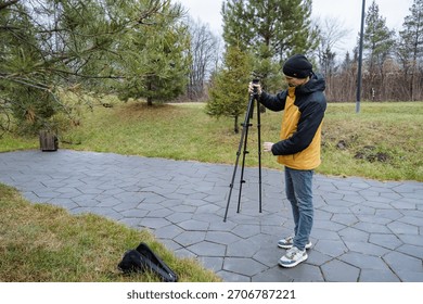 videographer assembling tripod on path adjusting mount for steady shot wearing yellow jacket and beanie camera attached nearby pavement and grass practical focused preparation for outdoor shoot