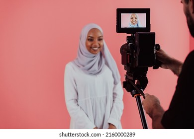 Videographer in digital studio recording video on professional camera by shooting female Muslim woman wearing hijab scarf plastic pink background. - Powered by Shutterstock