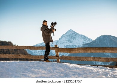 Videographer man shooting footage using camera mounted on gimbal stabilizer equipment. Snow and mountains in background.