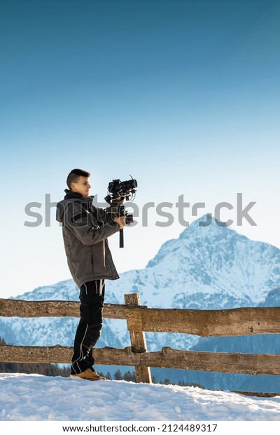 Videographer\
man shooting footage using camera mounted on gimbal stabilizer\
equipment. Snow and mountains in\
background.