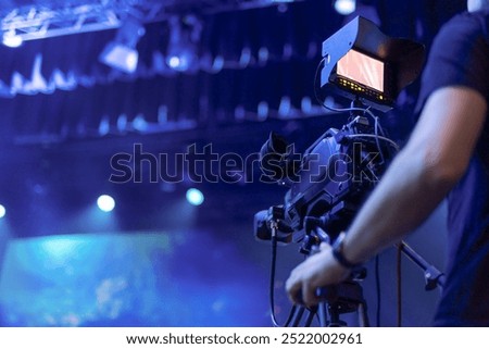 A videographer operates a professional camera during a live event, with vibrant blue stage lighting and blurred background creating a dynamic atmosphere. The camera screen displays the ongoing