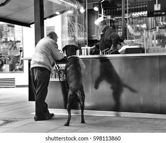 VIENNA, AUSTRIA - APRIL, 2011: Man With A Big Black Dog Standing At The Street Food Kiosk Waiting For A Hot Dog. Funny Shadow Of The Dog. Black And White Photo, Film Effect