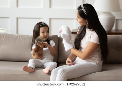 Vietnamese loving mother and toddler daughter wearing crowns casual clothes sit on couch playing with hedgehog and hare animal hand puppet toys. Educational games, having fun with kid at home concept