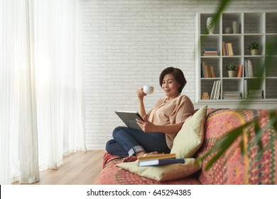 Vietnamese Senior Woman Drinking Tea And Reading Book On Tablet Computer