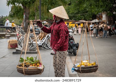 Vietnamese woman carrying baskets of fruits. Local woman vendor sells fruit that she carries in baskets at an old town Hoi An, Vietnam. Vietnamese street vendor woman carrying her goods in two baskets - Powered by Shutterstock