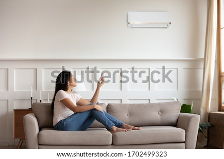 Vietnamese young woman relaxing on comfy couch in contemporary living room with air conditioner, holding remote control turning on or off cooler system, setting comfortable temperature at modern home