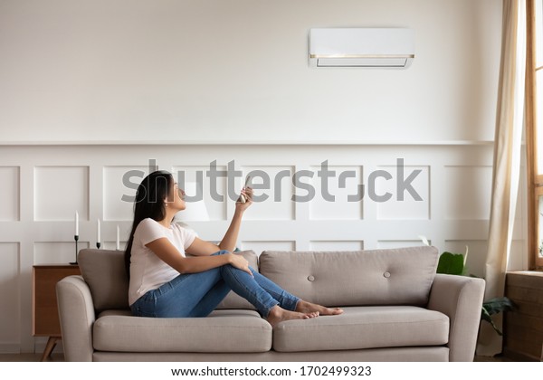 Vietnamese young woman relaxing on comfy couch in\
contemporary living room with air conditioner, holding remote\
control turning on or off cooler system, setting comfortable\
temperature at modern\
home