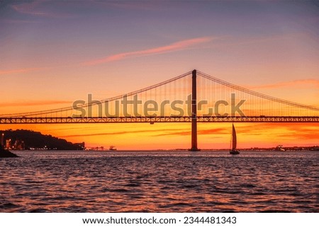 View of 25 de Abril Bridge famous tourist landmark of Lisbon connecting Lisboa and Almada over Tagus river with tourist yacht silhouette at sunset. Lisbon, Portugal