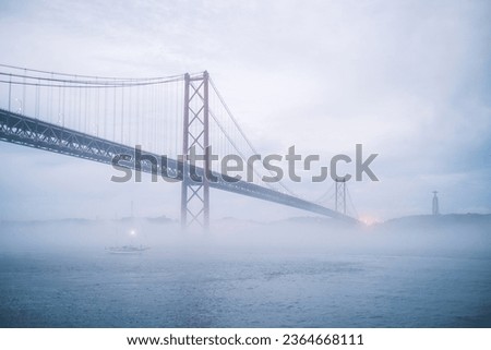 View of 25 de Abril Bridge famous tourist landmark of Lisbon connecting Lisboa and Almada in heavy fog mist wtih yacht boats passing under. Lisbon, Portugal
