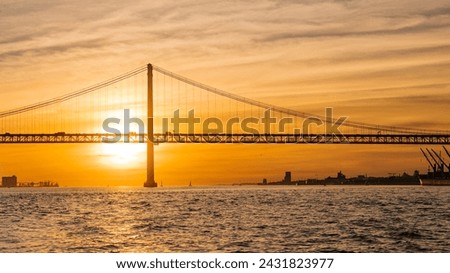 View of 25 de Abril Bridge famous tourist landmark of Lisbon connecting Lisboa and Almada over Tagus river with tourist yacht silhouette at sunset. Lisbon, Portugal