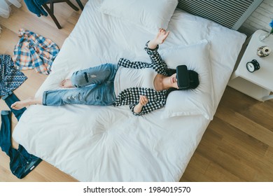 view from above carefree asian girl in casual wear is lying on bed having fun using vr headset in the bedroom with scattered clothing on floor at home.