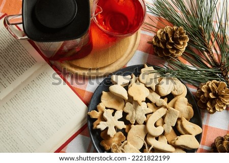 View from above a Christmas food composition with gingerbread pastries, teapot with herbal tea and open book, next to pine cones and fir-tree branch on kitchen table. Christmastime. Festive atmosphere