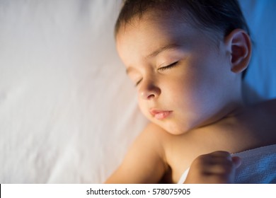 View From Above Close-up Portrait Of Adorable Baby Girl Sleeping In White Bed At Night. Little Toddler Taking A Nap In Dark Room In Lamplight. Bed Time For Kids. Bedroom And Nursery Interior.