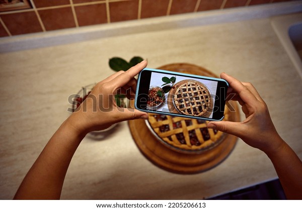View from above of the hands of a female food\
blogger holding a smartphone and taking a picture of a classic\
American puff pastry cherry pie. Mobile phone in live view mode.\
Baking pastry items