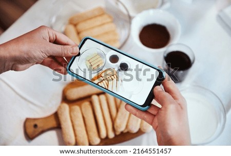 View from above. Hands using smartphone camera and taking photo of ingredients for Tiramisu dessert on the white tablecloth background. Mobile phone in live view mode