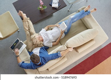 View From Above Of A Happy Couple Relaxing On The Sofa Together. Beautiful Woman Reading Book And Casual Man Using His Laptop While Lying On Their Cosy Couch At Home.