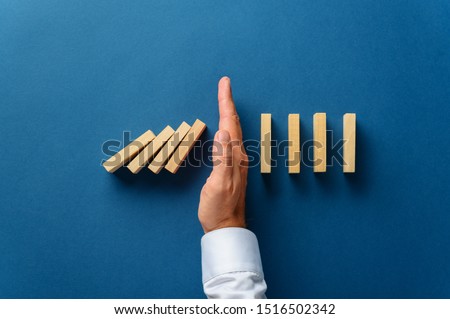 View from above of male hand interfering collapsing dominos in a conceptual image of business crisis management. Over navy blue background.