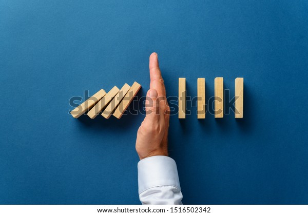 View from above of male hand interfering\
collapsing dominos in a conceptual image of business crisis\
management. Over navy blue\
background.