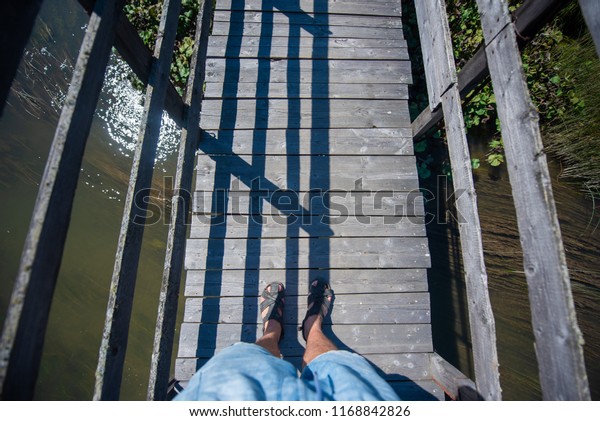 view from above on the man\'s feet in sandals. feet\
of a man standing on an old wooden bridge. the man is standing on\
the bridge over the river