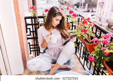 View from above pretty girl in pajama having breakfast on balcony in the morning in city. She holds a cup, reading on tablet