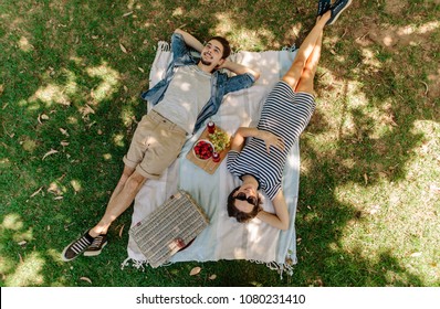 Vista desde arriba de un hombre y una mujer relajados tendidos en el parque con cesta de mano, aperitivos apetitosos y cervezas en manta. Pareja de picnic echada en una manta en el césped al aire libre. Foto de stock