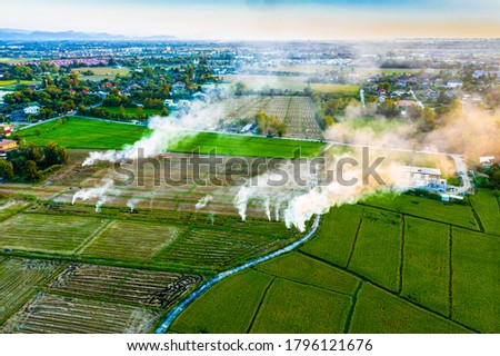 View from above of rice fields with small fire burning spots and smoke of the countryside in a blur background of town and village from afar in a evening of Chiengmai, Thailand