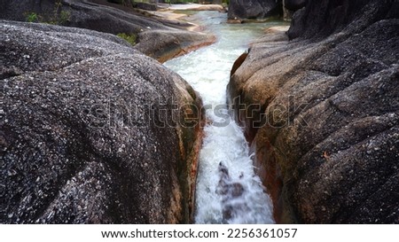 View From Above, A River Of Fresh Fresh Water Flowing From A Gap Between Two Large Black Mountain Rocks, In Daya Baru Village, During The Day