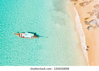 View from above, stunning aerial view of two people walking on a beautiful tropical beach with white sand, turquoise clear water and a traditional long-tail boat. Banana beach, Phuket, Thailand.