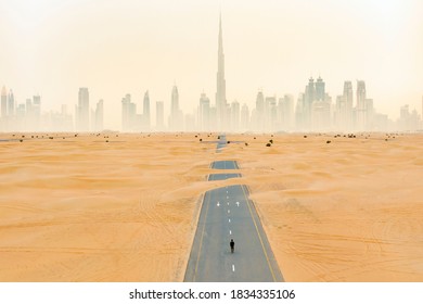 View from above, stunning aerial view of an unidentified person walking on a deserted road covered by sand dunes with the Dubai Skyline in the background. Dubai, United Arab Emirates. - Shutterstock ID 1834335106