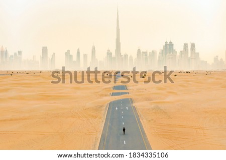 View from above, stunning aerial view of an unidentified person walking on a deserted road covered by sand dunes with the Dubai Skyline in the background. Dubai, United Arab Emirates.