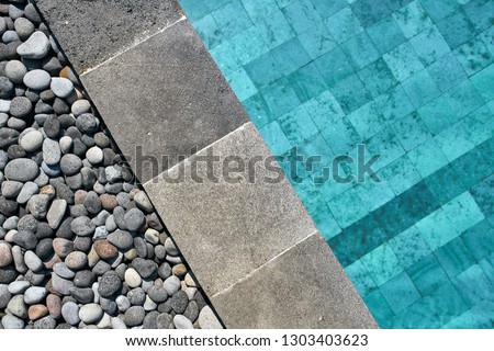 View from above at a swimming pool with blue water outdoors. It has a tiled gray edge which is decorated with pebbles. Closeup horizontal photo..