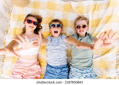 View From Above Of Three Kids In Eyeglasses Lying On Plaid Stretching Hands To Camera At Summer Beach. Childhood, Vacation, Summer Camp.