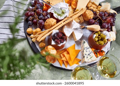 A view from above through the greenery of a large round Charcuterie Board and glasses of white wine on a green table