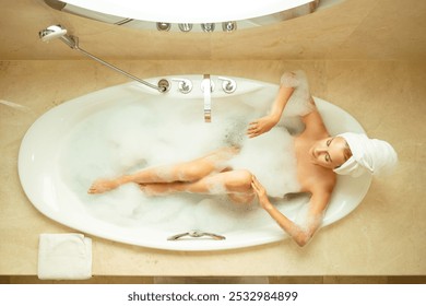 View above Woman with towel on her head taking hot relaxing bath with salt, bubble bath. Beautiful young woman enjoying bath. Lady on hotel vacation lying in bathtub. Girl relaxing in float bath. - Powered by Shutterstock