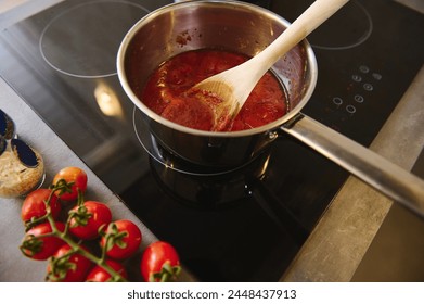View from above of a wooden spoon in a steel pan with boiling homemade sauces of ripe organic juicy red tomatoes. branch of fresh red tomato cherry on the kitchen counter near a black induction cooker