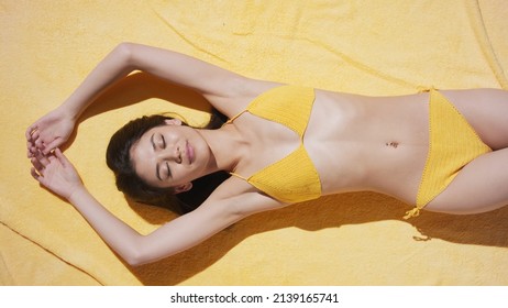 View From Above Of Young Attractive Slim Asian Brown-haired Woman In Yellow Crochet Bikini Lying On Yellow Beach Blanket Puts Her Hands Up Sunbathing And Smiling | Sun Cream Commercial