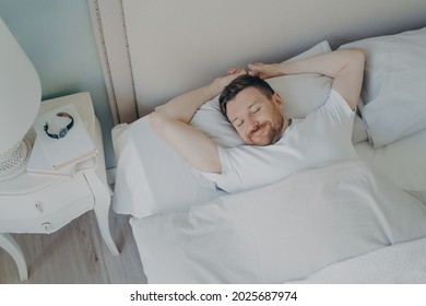 View From Above Of Young Happy Relaxed Caucasian Man With Attractive Smile Sleeping, Lying In Cozy Bed With White Beddings. Healthy Care Good Night Sleep And Rest Concept