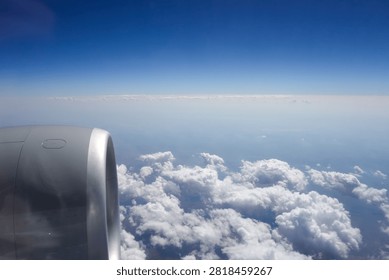 View of an airplane engine from a passenger window during flight above a layer of white clouds. Aerial travel scene featuring cloud formations, blue sky, and glimpses of the landscape through breaks in the clouds.