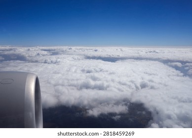 View of an airplane engine from a passenger window during flight above a layer of white clouds. Aerial travel scene featuring cloud formations, blue sky, and glimpses of the landscape through breaks in the clouds.