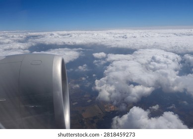 View of an airplane engine from a passenger window during flight above a layer of white clouds. Aerial travel scene featuring cloud formations, blue sky, and glimpses of the landscape through breaks in the clouds.