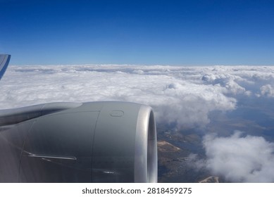 View of an airplane engine from a passenger window during flight above a layer of white clouds. Aerial travel scene featuring cloud formations, blue sky, and glimpses of the landscape through breaks in the clouds.