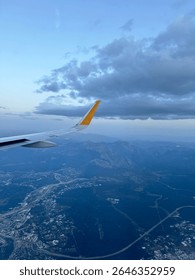 View from airplane window of wing flying over city and mountains under cloudy sky. Feeling of travel, introspection, and human connection to vast landscapes, captured in a moody, cinematic atmosphere