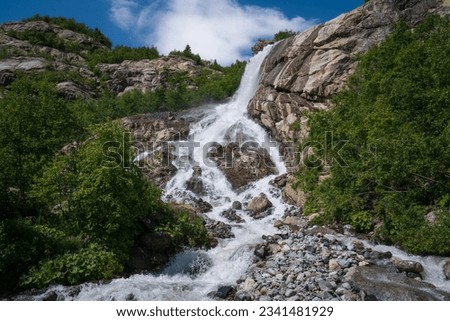 View of the Alibek waterfall formed by the fall of the Jalovchatka River from the Alibek glacier on a sunny summer day, Dombay, Karachay-Cherkessia, Russia