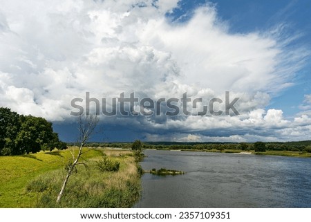 View along German-Polish border river Oder to big thunderstorm front from which partly rain falls, sunny weather, green river bank with tree, river bend, Germany Poland border area, Oderbruch