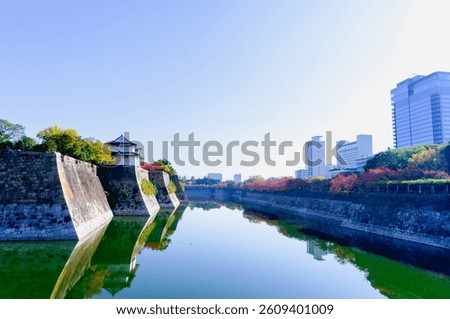 View along outer moat and wall of Osaka Castle with buildings and park in morning of colorful autumn season or fall foliage, Japan. Cityscape. Water reflection mirror.