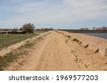 A view along the top of the Lake Jackson State Park dam road from a high perspective showing water of the lake.  Dry road, masonry walls and green fields with trees and a light blue sky.