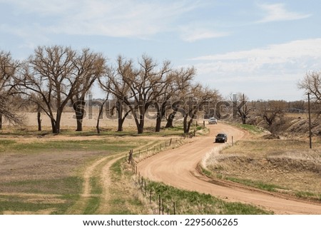 A view along the top of the Lake Jackson State Park dam road from a high perspective showing car driving down dirt road.  Dry road, trees with buds and green fields with a light blue sky.