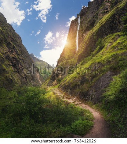 View with amazing Himalayan mountains covered green grass, high waterfall, beautiful path, fgreen trees, blue sky with sun and clouds in Nepal at sunset. Mountain canyon. Travel in Himalayas.Landscape